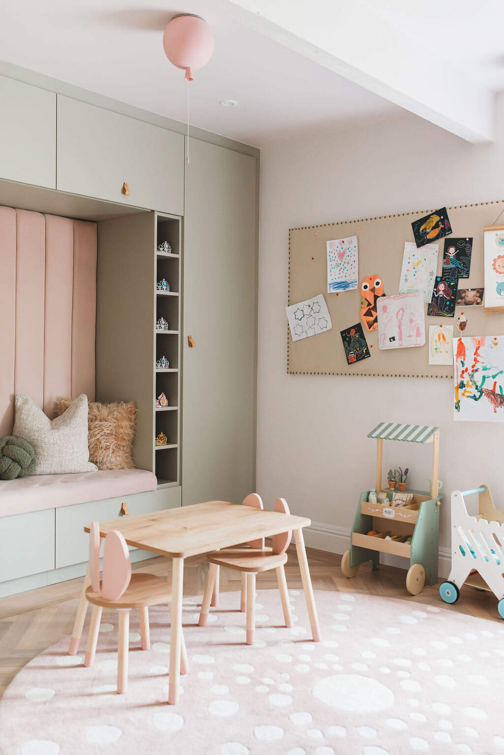 A cozy children's playroom with pastel-colored decor. It features a built-in cushioned seating nook with shelves, a wooden table with pink chairs, a board displaying children's artwork, a toy cart, and a play market stand. A pink balloon hangs from the ceiling.