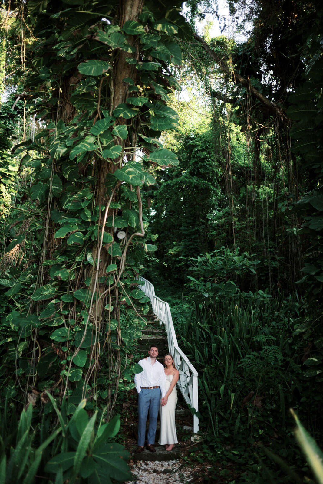 The engaged couple is standing at the foot of a very high stone staircase in the middle of lush greens in Jamaica.