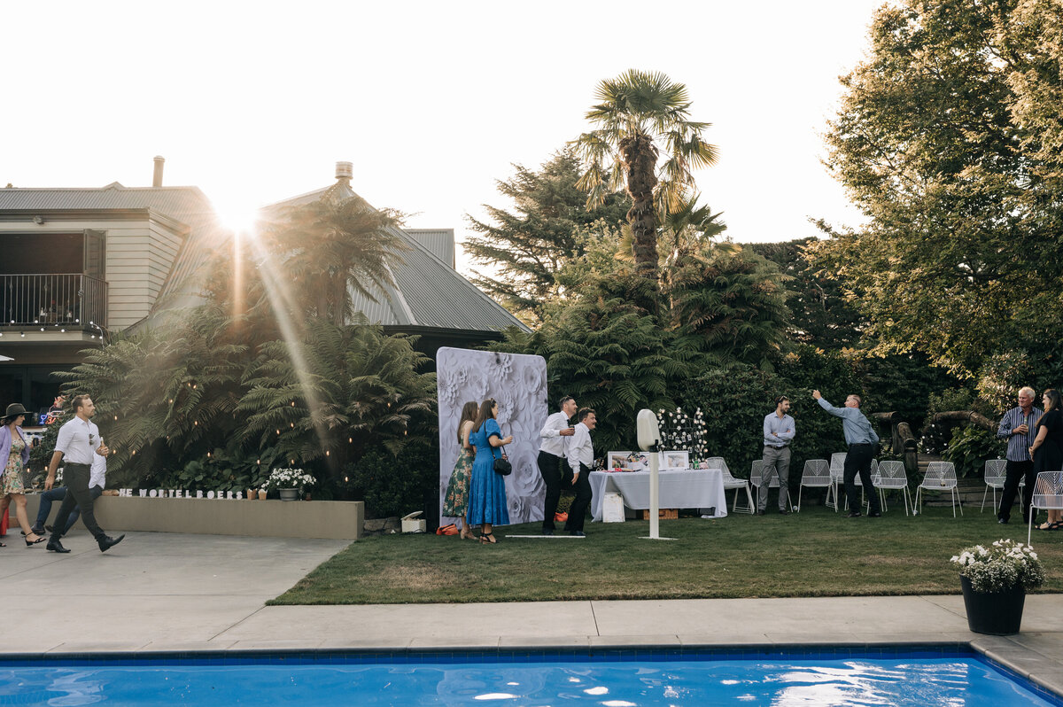 sun sets over a wedding reception on a lawn beside a pool while guests use the photo booth near christchurch