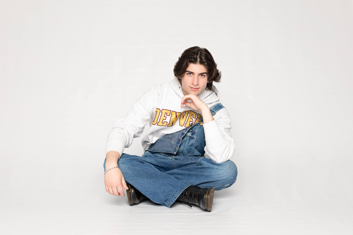 A person with long dark hair sits cross-legged on the floor against a plain white background. They are wearing a white Denver hoodie and blue denim overalls, resting their chin on one hand while looking at the camera.