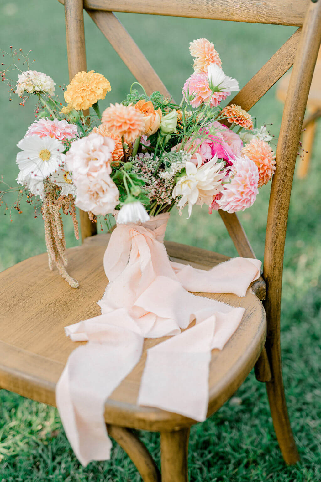Colorful wedding ceremony flowers with trailing ribbon tied to wooden chair on lawn.