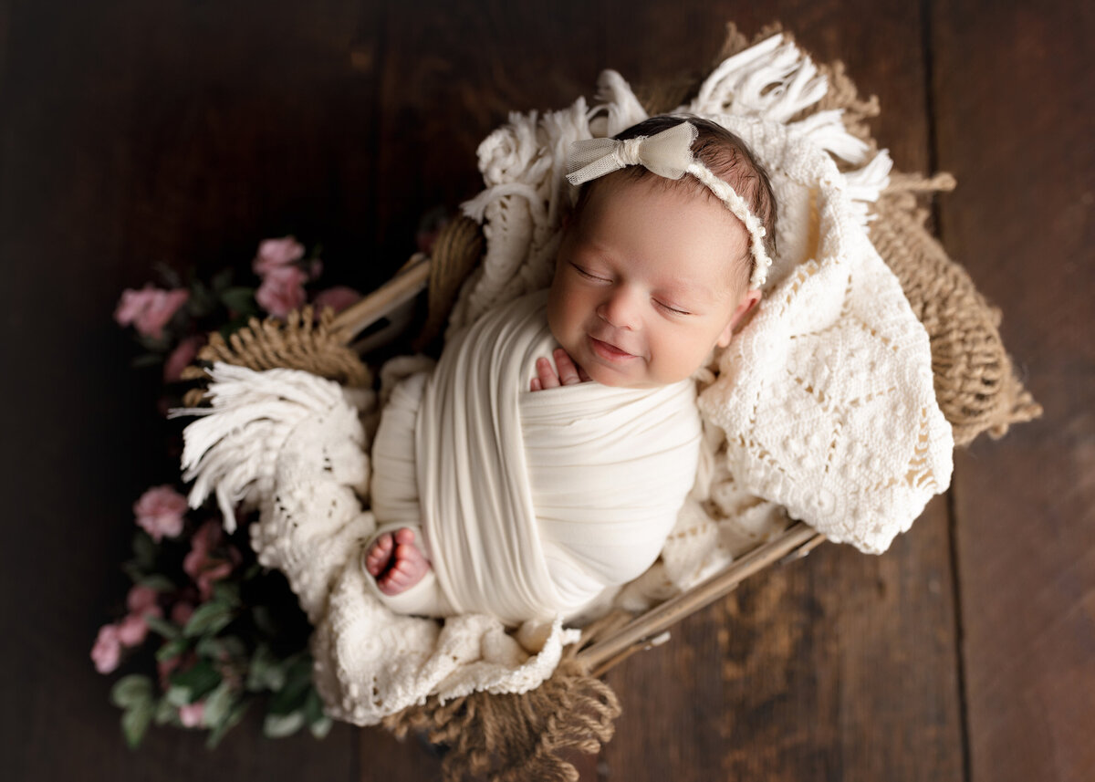 Baby girl sleeping in wood bassinet for newborn photoshoot. Aerial image. Baby's fingers and toes are peeking out of the wrap. Baby is resting atop of a crochet blanket with blurred flowers below the bassinet.