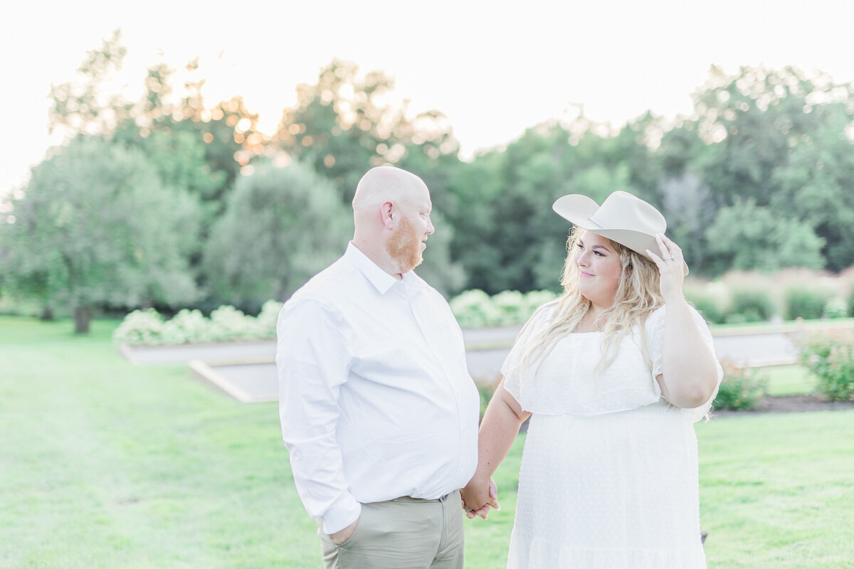 Couple looking at each other holding hands both wearing white. The soon to be bride is wearing a cowboy hat