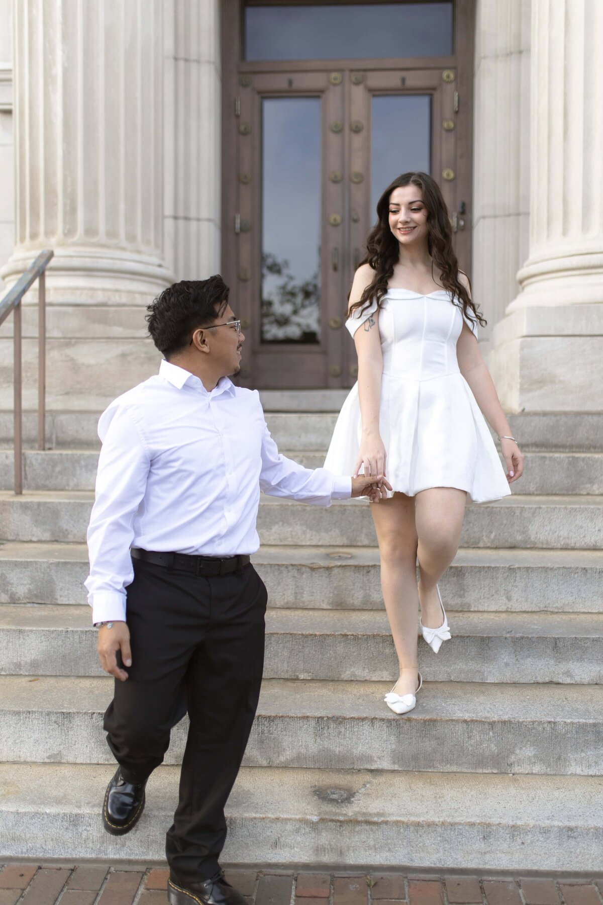A couple shares a sweet moment as the groom-to-be helps his fiancée down the steps of a historic courthouse building in Jacksonville, Florida. The bride-to-be wears a chic off-the-shoulder white mini dress and heels, while her partner is dressed in a white shirt and dark trousers. The grand columns and architecture add a timeless elegance to this modern love story. Captured by Heartful Films, a Jacksonville engagement photographer and videographer known for capturing romantic, stylish moments with personality.