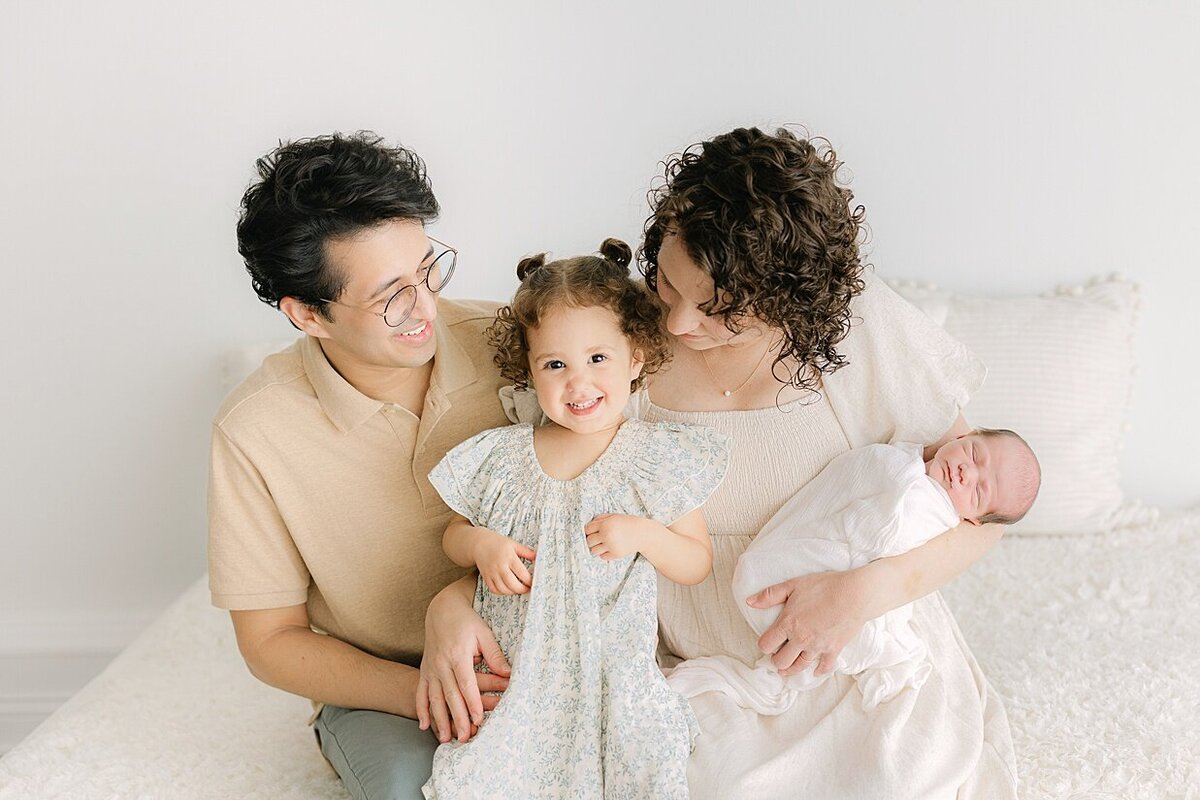 Family of 4 sits on a bed dressed in creams and sage with mom and dad smiling at their toddler daughter as she giggles while Mom holds her baby during their session with an Indianapolis newborn photographer