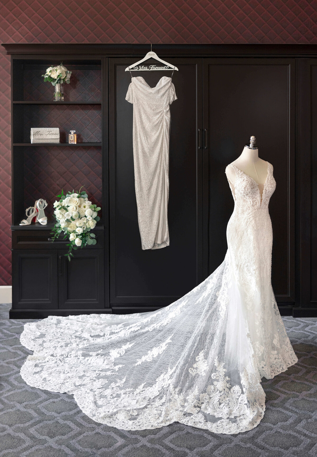 A long, elegant white lace wedding dress with a train is displayed on a mannequin. A sparkly silver dress hangs on a wardrobe behind it. On a shelf are a bouquet, a pair of white heels, and a Mrs. Forever sign.