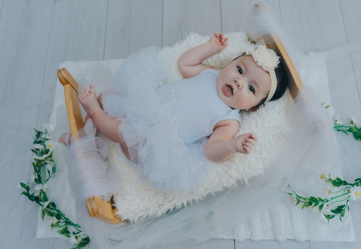 Baby girl lying on a fluffy peach blanket, wearing a white bow headband and a pale pink outfit, smiling softly at the camera.