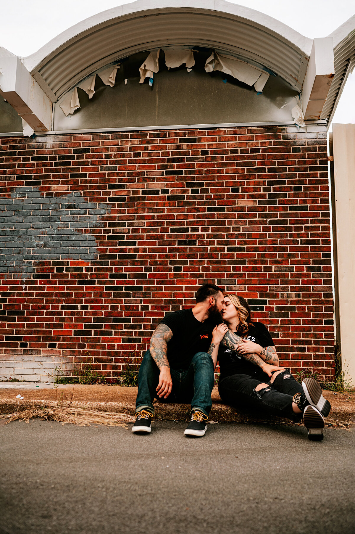 tattooed couple sitting on ground in urban location, woman is leaning in to passionately kiss male partner.