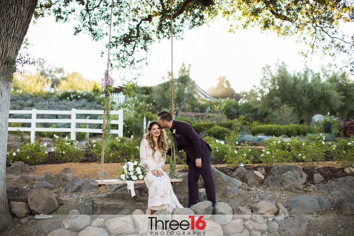 Bride smiles while sitting on a swing bench as her Groom kisses the top of her head