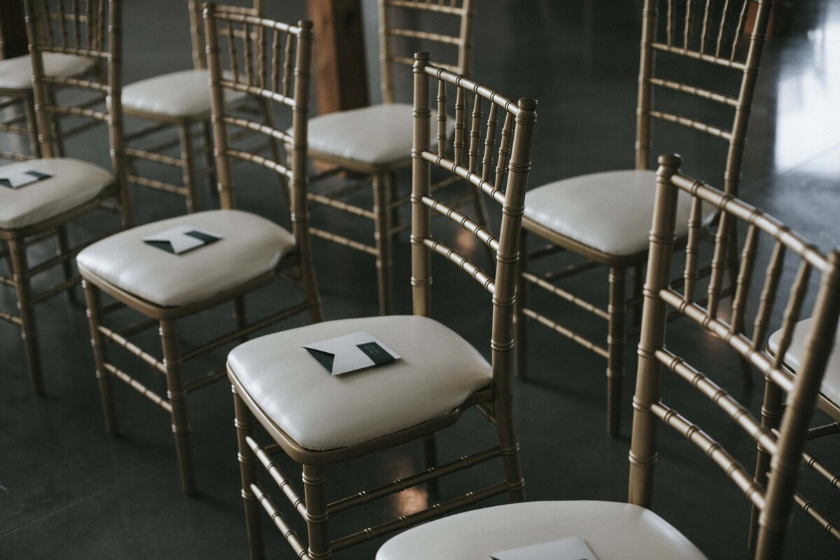 White and dark green wedding place cards set atop gold chairs with ivory seats.