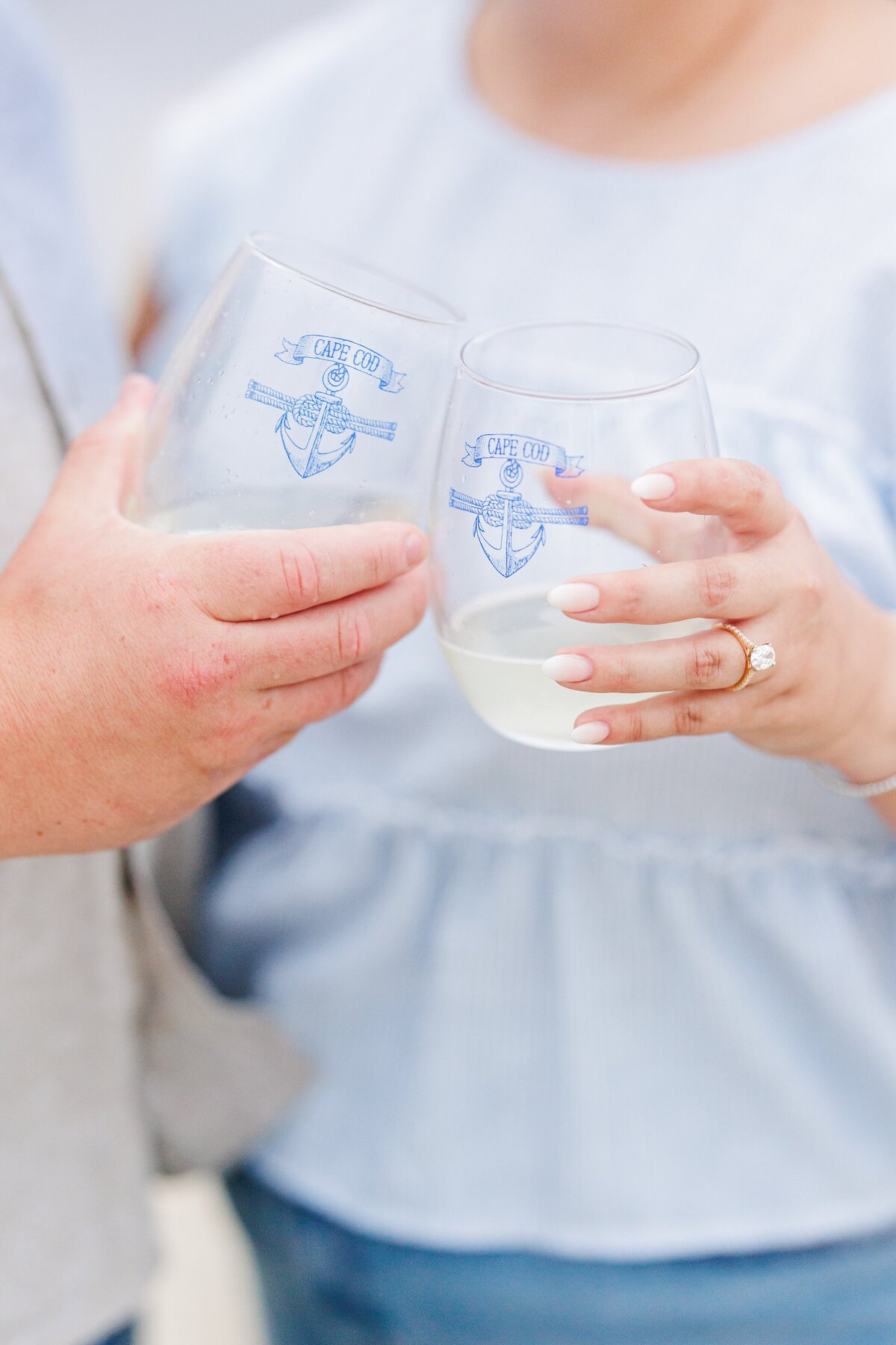 Close-up of two stemless glasses with a blue “Cape Cod” anchor design, held by a couple toasting with white wine, the woman’s engagement ring visible, at Chatham Bars Inn in Chatham, Massachusetts — representing the portfolio of a Cape Cod engagement photographer.