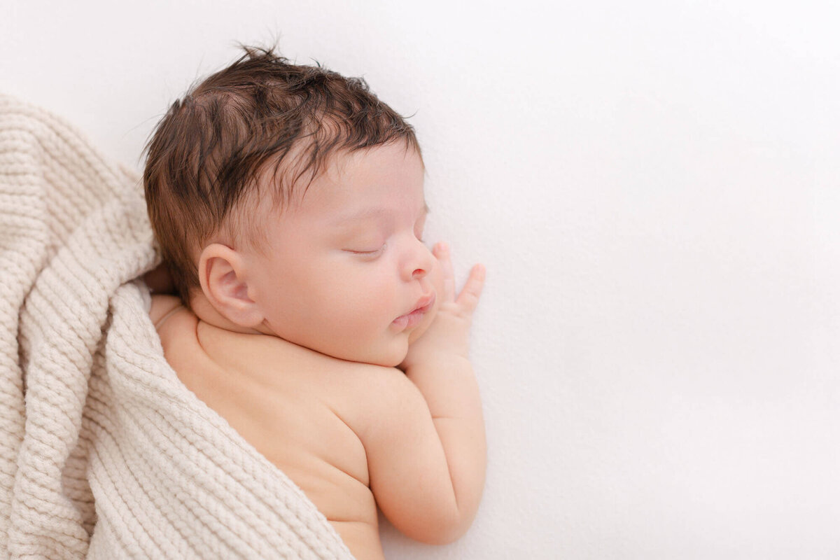 Newborn baby sleeping while laying on stomach with a beige textured blanket draped over his back.