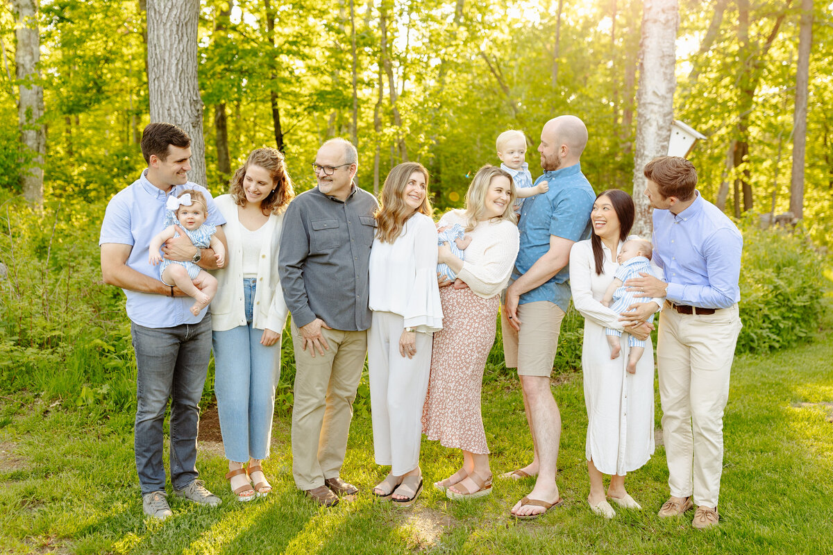 A group of ten people, including adults and infants, stands outdoors in a sunlit forest setting. Captured by a Brainerd Lakes Area Photographer, they are smiling and dressed in casual, light-colored clothing. Some adults are holding babies against the backdrop of lush trees and grass.