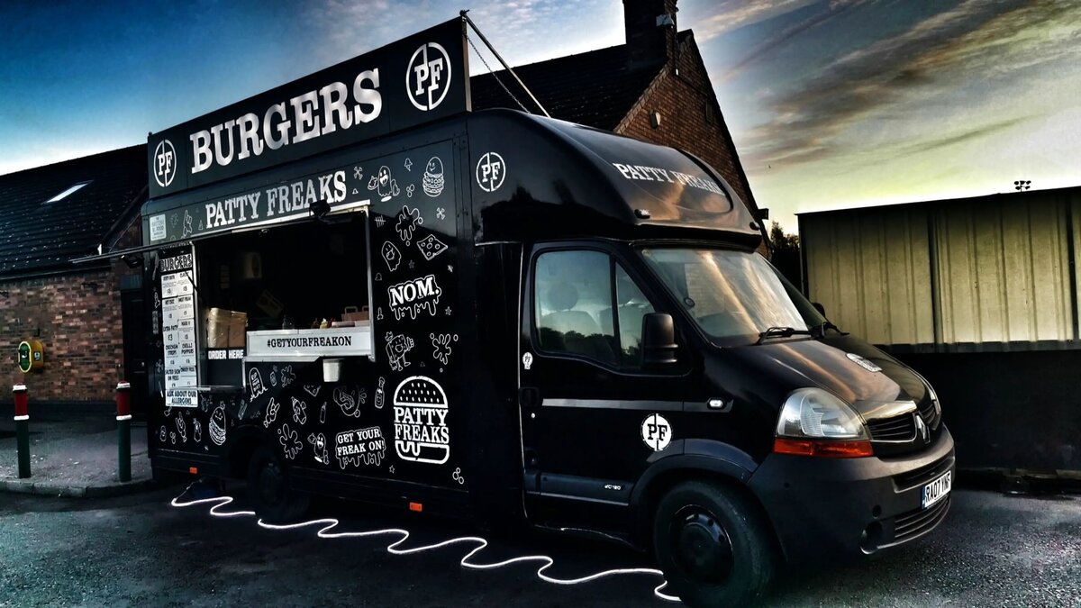 The Patty Freaks Burger Food Truck and mobile catering van. Black with white writing, parked outside a building with the sun setting.