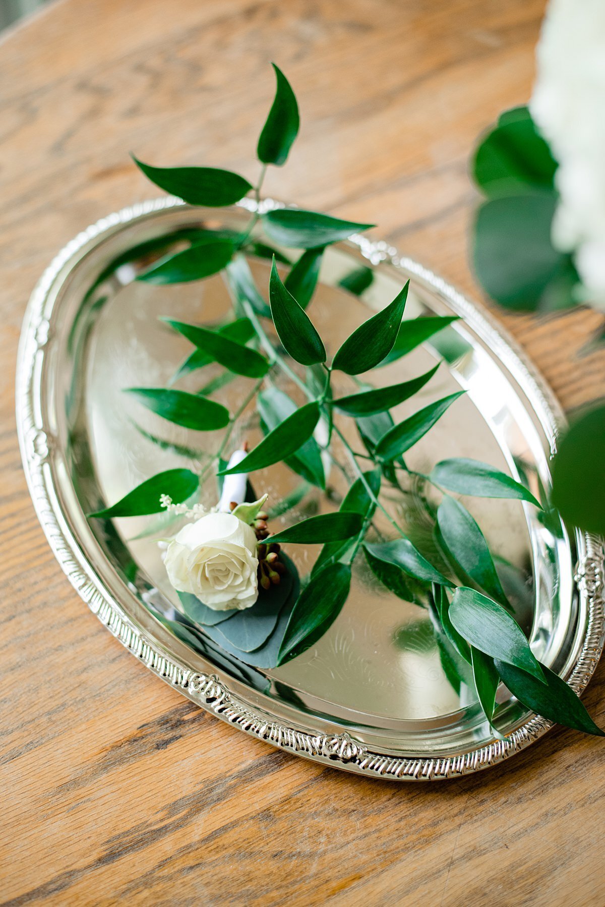 The groom's boutonniere sits on a small sliver tray with accenting greenery. The silver platter is placed on a wooden table.