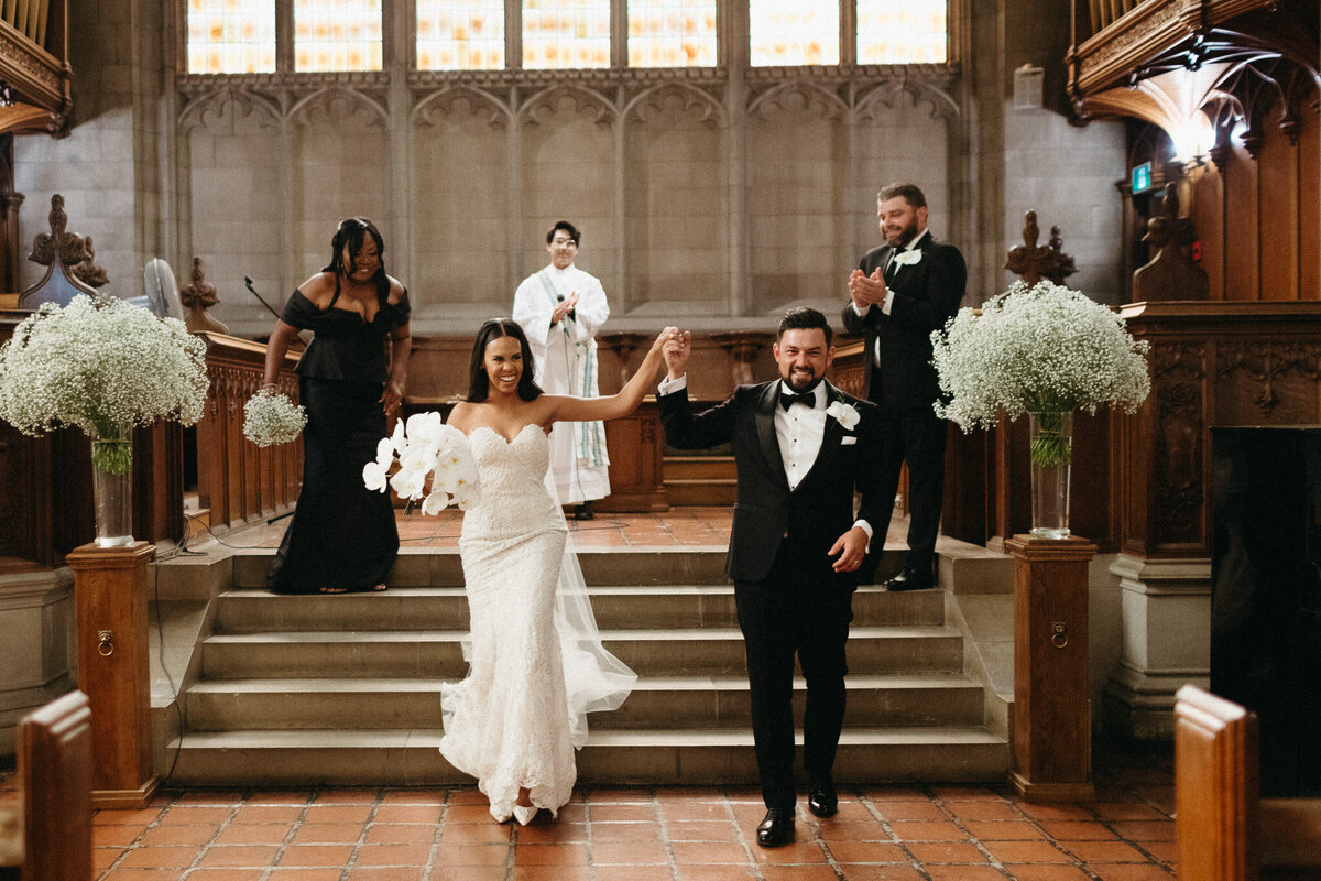 A bride and groom holding hands walk down the aisle in a church, followed by bridesmaids and groomsmen. Captured in perfect harmony by top wedding photographer in Toronto, Jennifer van Son, the couple radiates joy as they smile in their elegant formal attire.