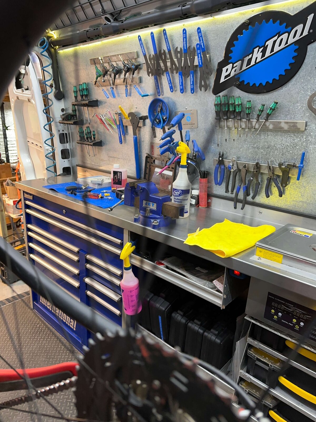 A well-organized bike repair station inside a van, featuring an array of tools on a wall-mounted board, a workbench with cleaning supplies, and storage drawers. A bicycle wheel is visible in the foreground