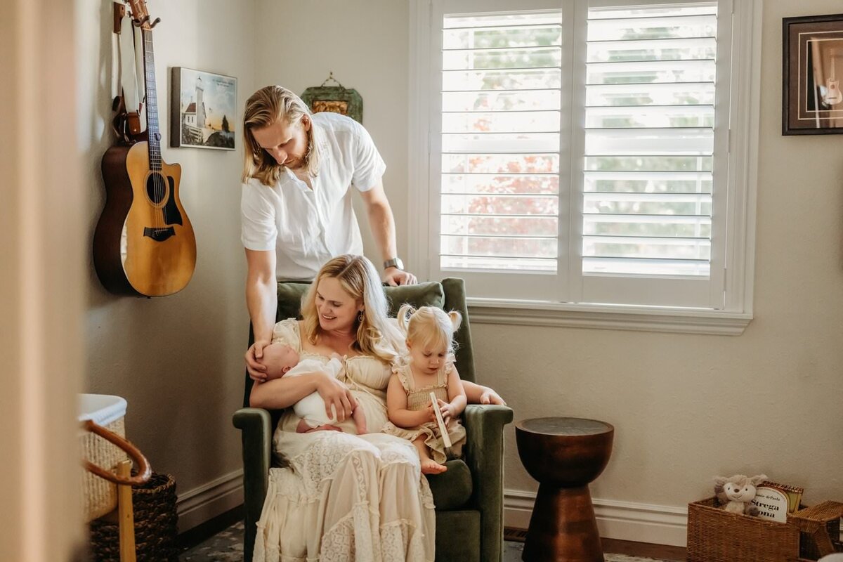 mom holds toddler and newborn son in their nursery. dad looks down at them. toddler reads a book