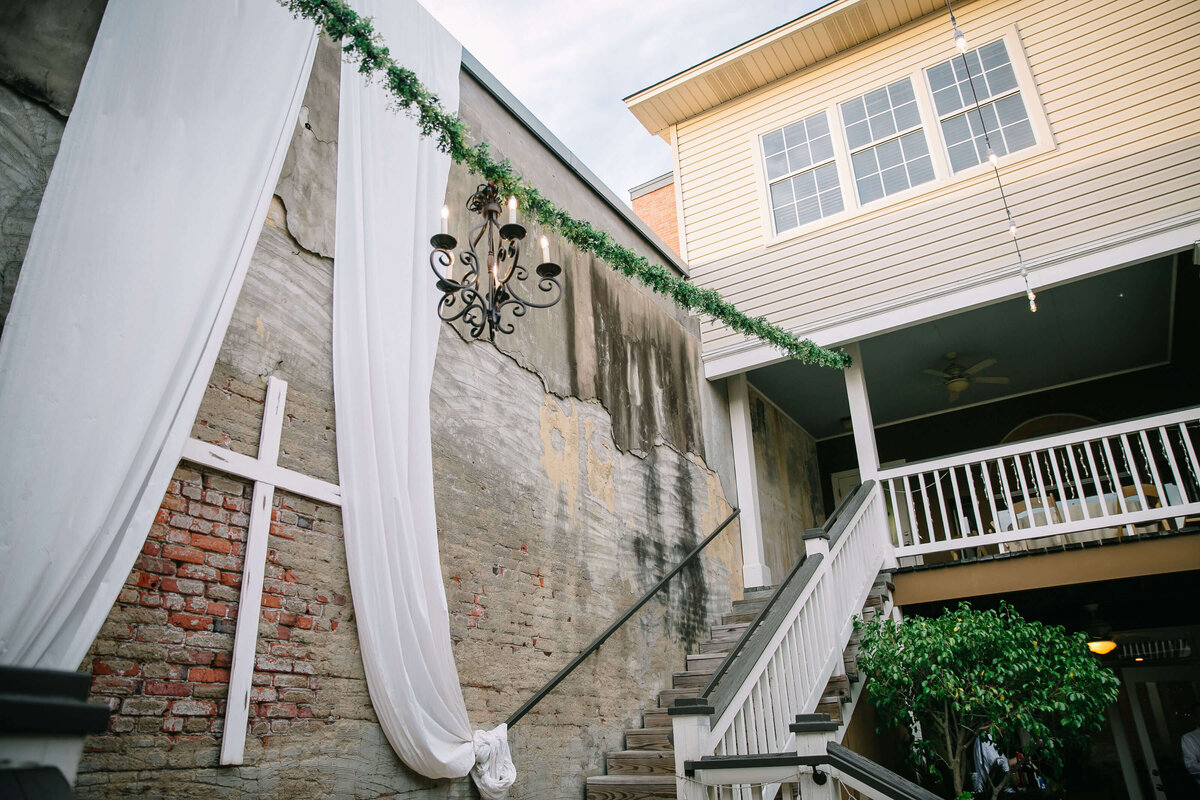 An outdoor staircase is decorated with white drapery and a cross against a weathered brick wall. A chandelier hangs above, and green garland borders the railing.