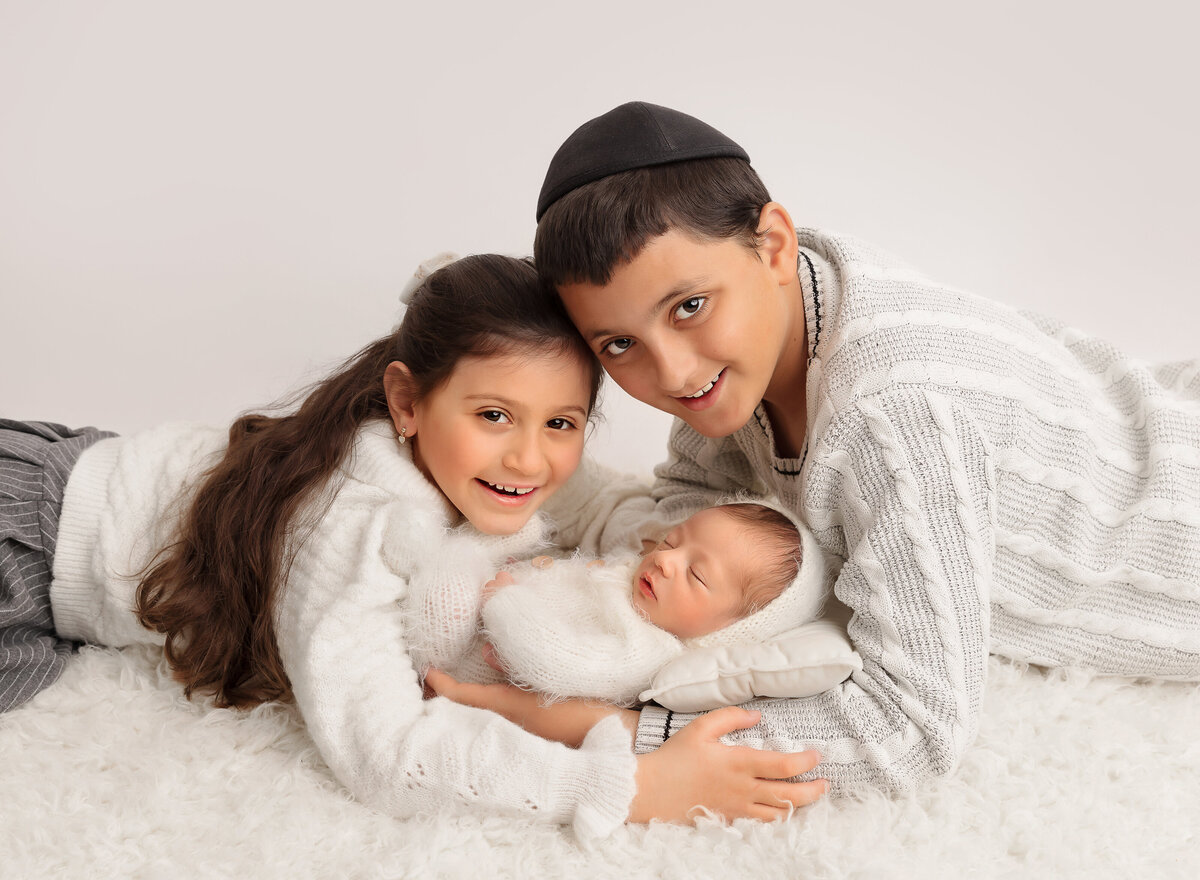 Big brother and big sister are laying on the ground facing each other with their newborn baby brother held between them. Brother and sister are smiling at the camera. Captured by top Brooklyn newborn photographer Chaya Bornstein
