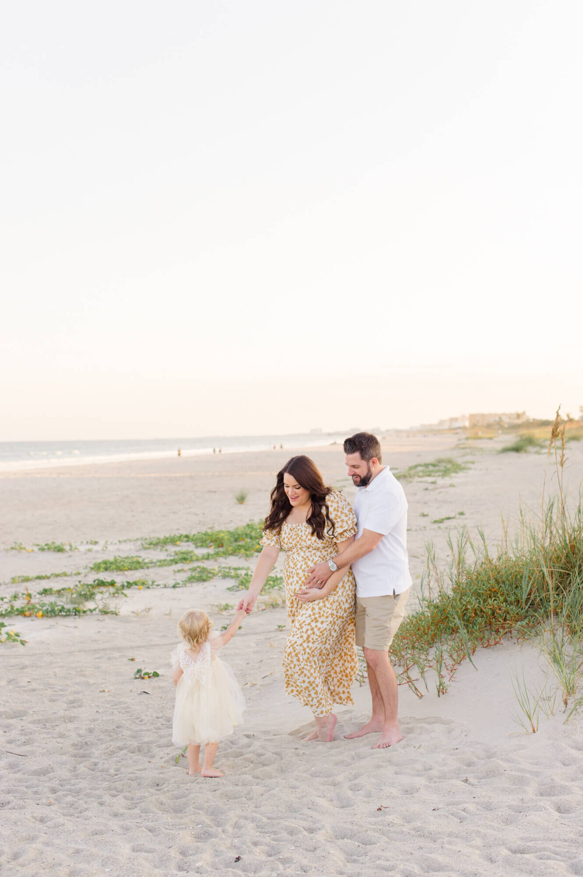 New parents stand on the beach near the dunes watching their young daughter hand them shells at the end of their Orlando maternity photographer session