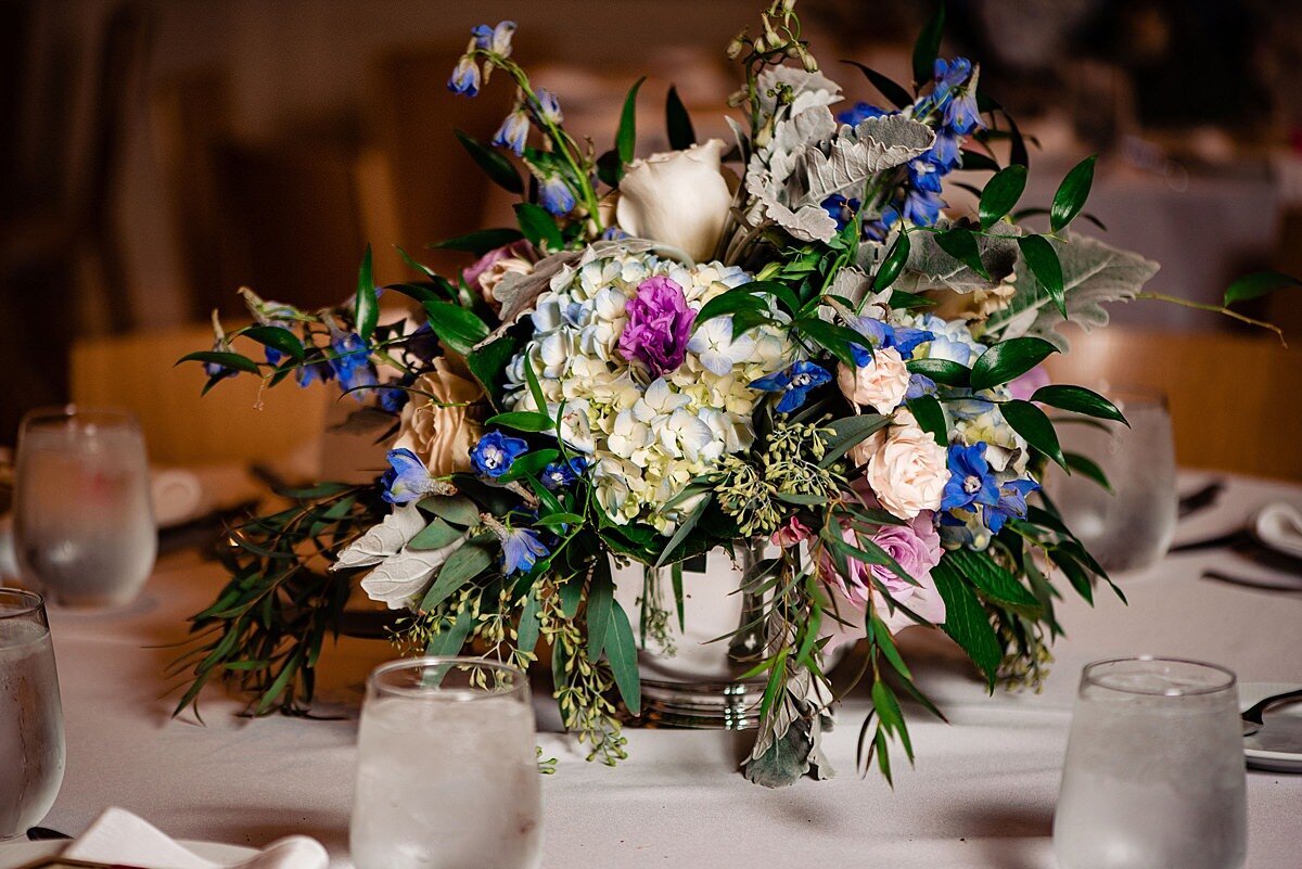 White hydrangea centerpiece in a silver footed bowl with blue and pink floral accents with greenery sitting on a table with a white linen and glasses filled with ice water at a wedding in Nashville, TN.