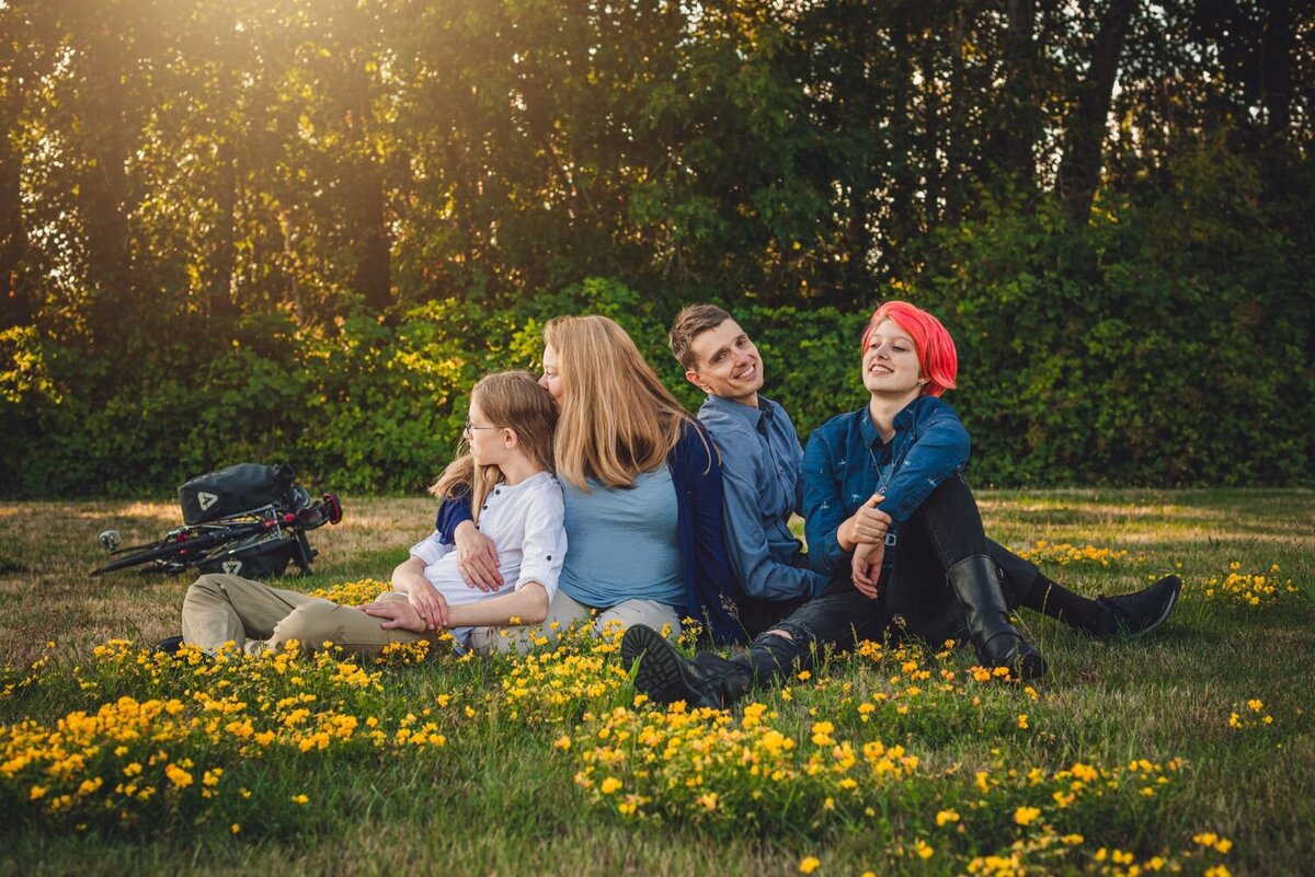 The image shows four people sitting on grass with yellow flowers. They are sitting back-to-back in a row. From left to right: a person with long blonde hair and glasses wearing a white shirt and beige pants; a person with long blonde hair wearing a blue shirt and a dark blue cardigan; a person with short brown hair wearing a blue button-up shirt; and a person with short pink hair wearing a dark blue shirt and black pants with black boots. In the background, there are green bushes and trees. A bicycle with a black bag is lying on the grass to the left. The lighting suggests it is late afternoon or early evening.