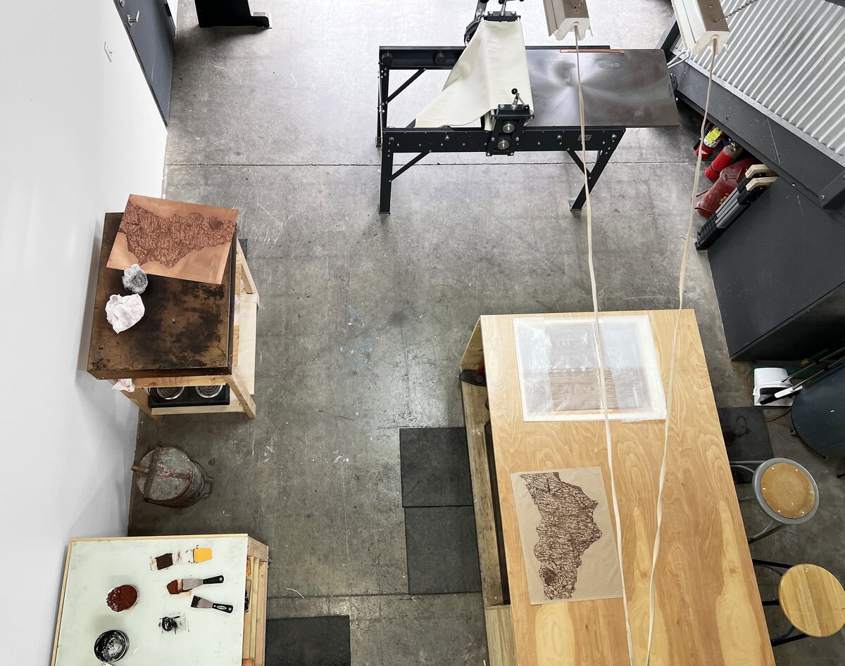 Overhead view of a printmaking workspace with a copper plate featuring an etched design, ink slabs, and a rolling press.