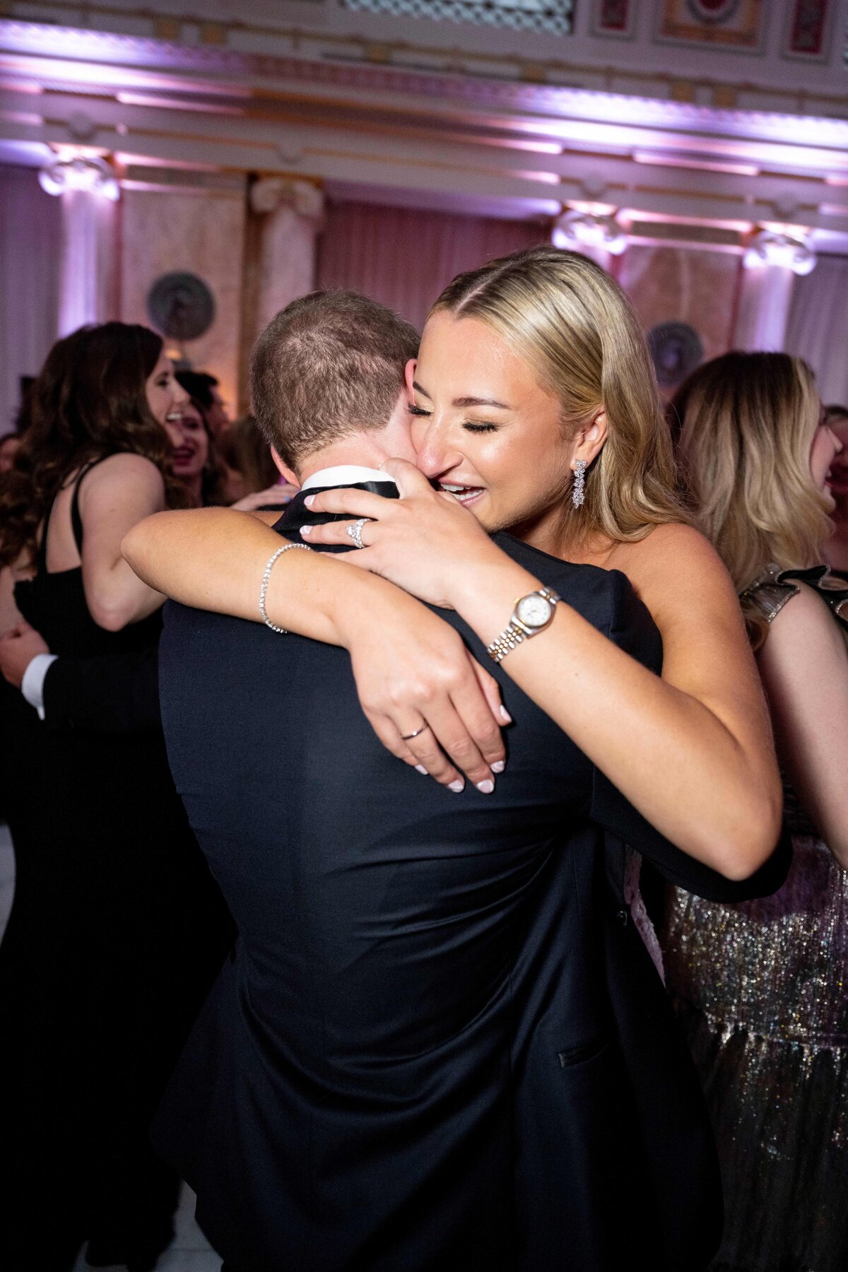 A woman with long blonde hair happily hugs a man in a suit at a formal event. The background shows other people dressed in formal attire, with elegant lighting and decor.