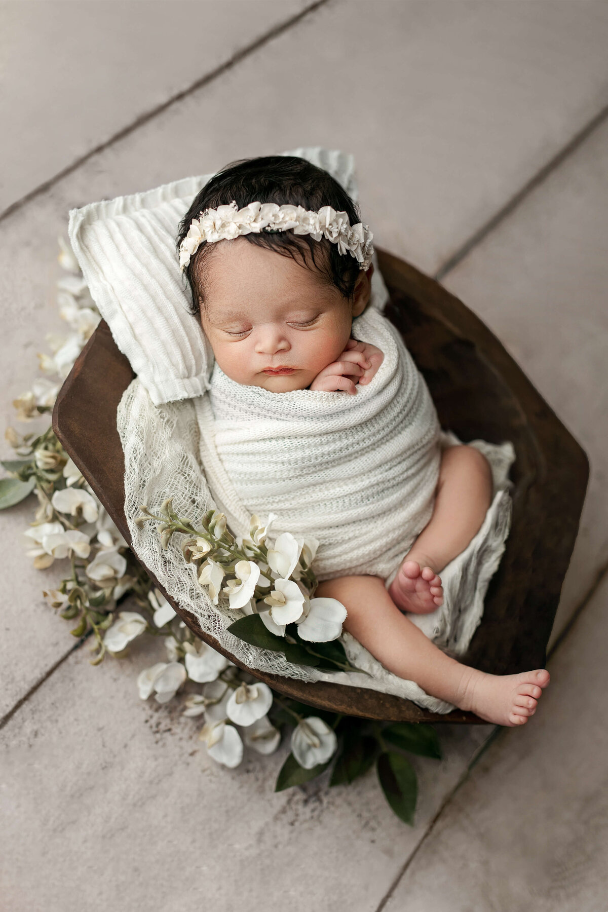 newborn baby girl in a white swaddle laying in a brown wooden bowl with flowers  on a white wood floor  with floral headband at a newborn photo shoot in Northern VA