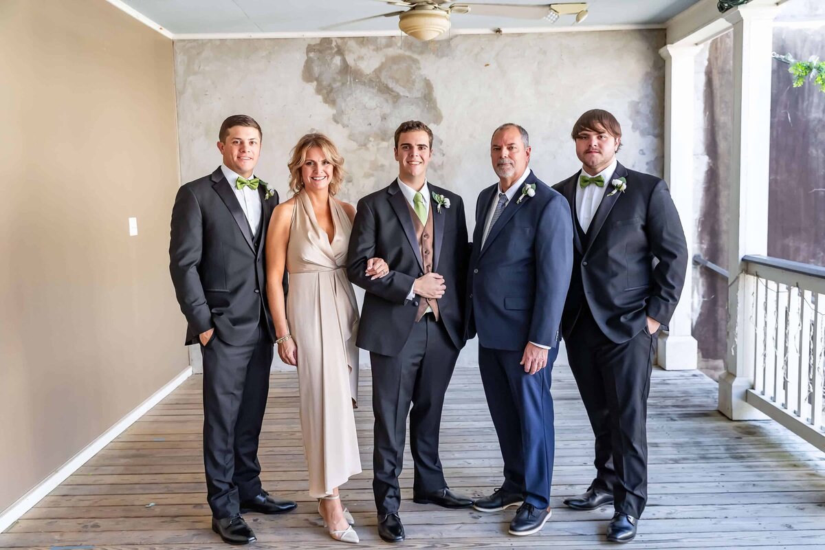 A group of five people dressed formally stand on a balcony with white railings. Three men in black suites, a woman in a beige dress, and a man in a navy suit smile at the camera.