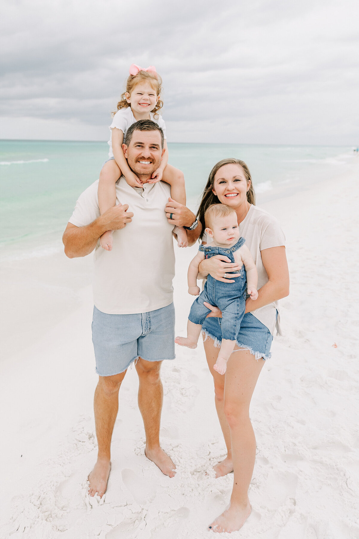 A happy family of four poses on the white sandy shores of Orange Beach, AL. The father, dressed in a neutral polo and denim shorts, carries his young daughter on his shoulders as she grins with a pink bow in her curly hair. The mother, smiling in a beige top and denim shorts, cradles a baby in denim overalls. The turquoise waves and soft clouds create a stunning backdrop for this cherished beach moment