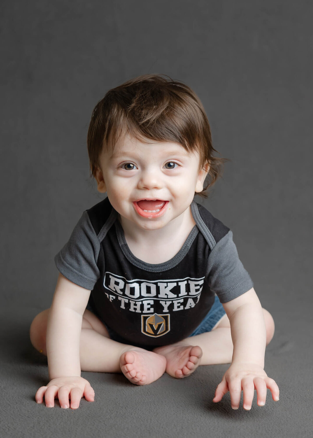 Happy infant boy  sitting up on a gray background with a Vegas Knights shirt on, smiling for his daycare portrait