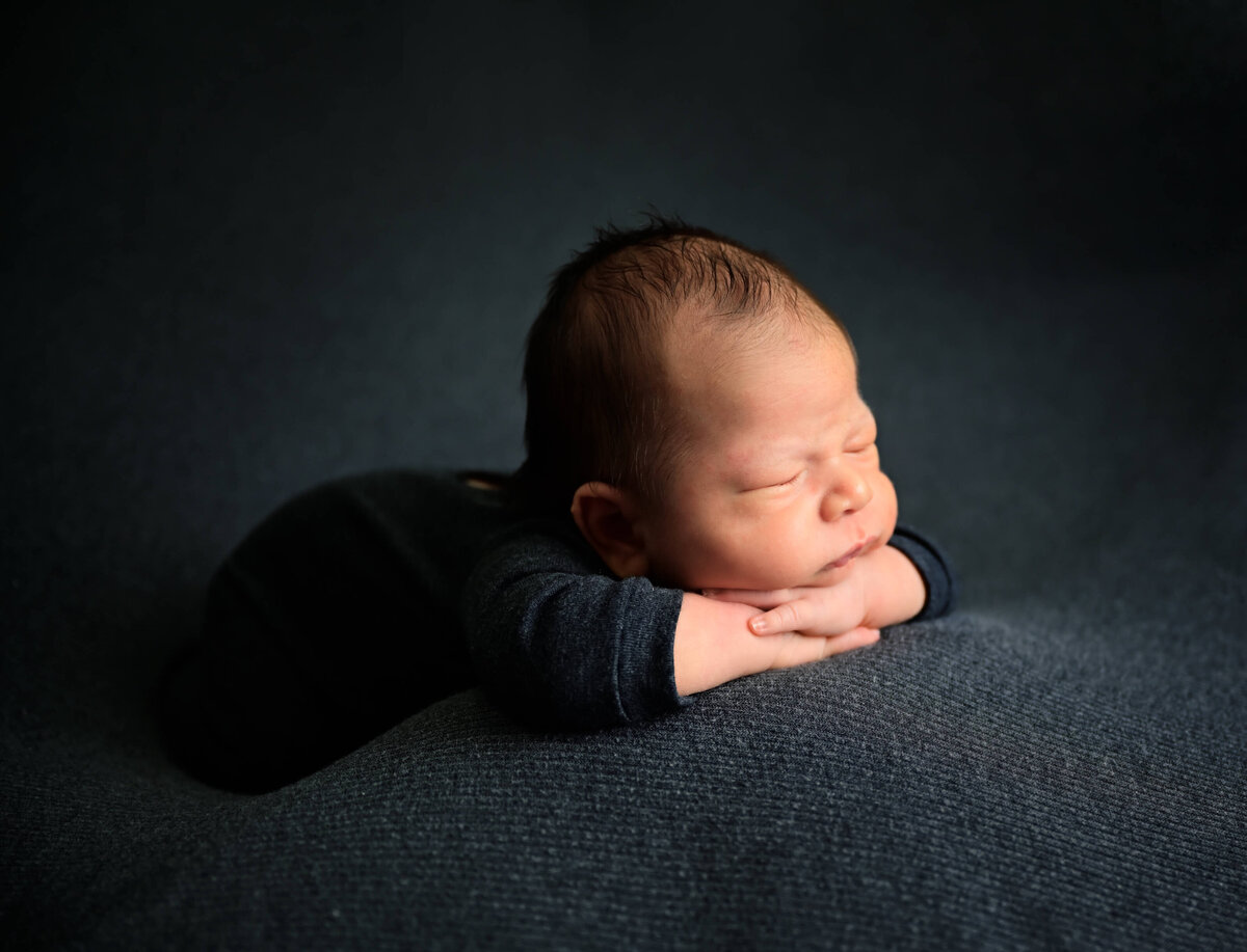 Side angle of newborn positioned on his tummy with his chin on his arms and hands. Baby is dressed in charcoal gray and on a matching blanket. Photographed by Beth Benecki of E. Benecki Photography.