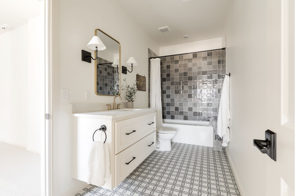 Bathroom with patterned black and white floor tile, creamy white vanity with brass faucet, arched mirror with dual sconces, and a tub surround clad in glossy charcoal zellige tile.