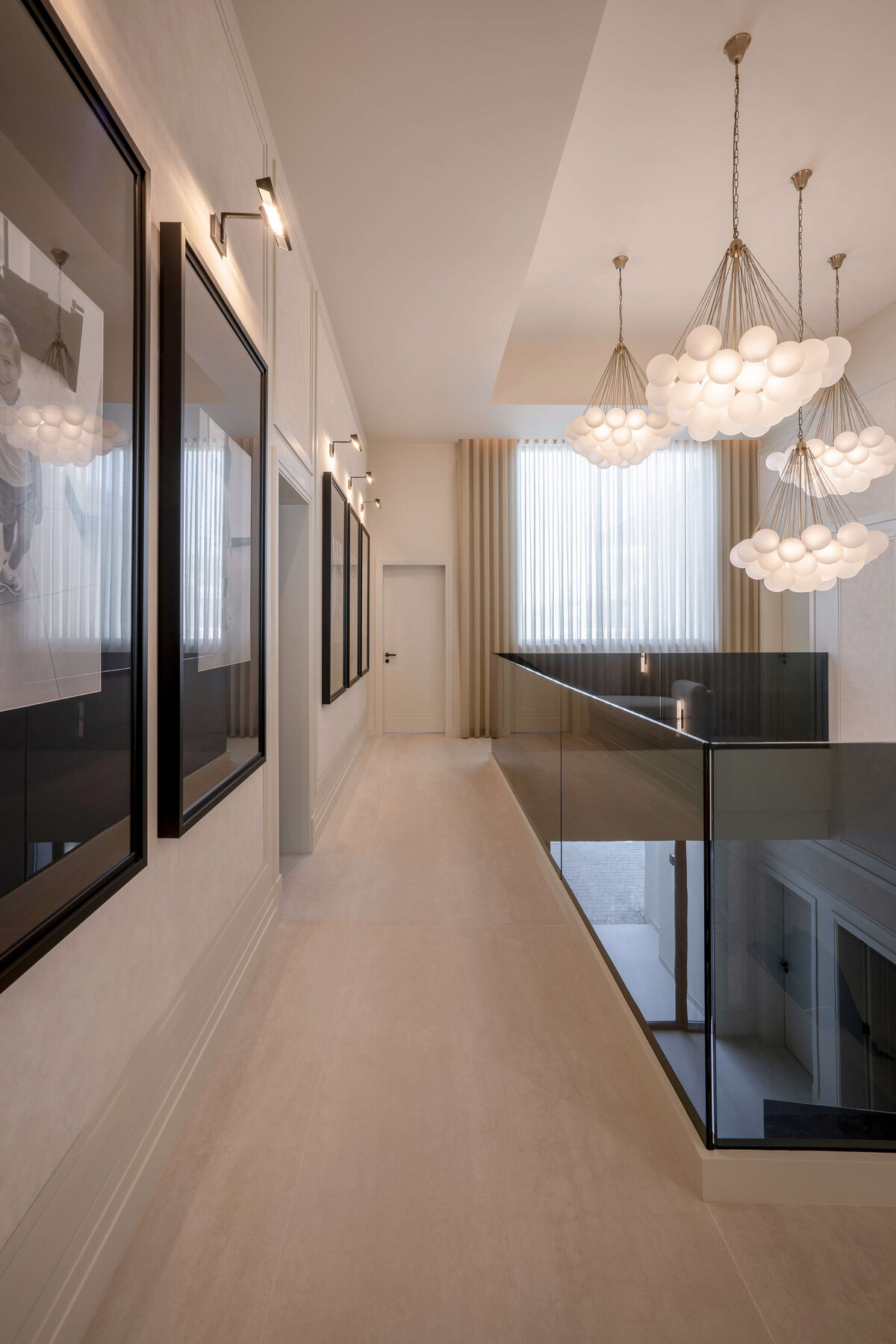 The top of a modern white staircase, bordered by a sleek, black glass railing on the right side. The staircase leads to a large, open landing, with the black glass railing continued through the hallway. Above, bold, bubbly chandeliers emit a soft light and add a contemporary style to the space.