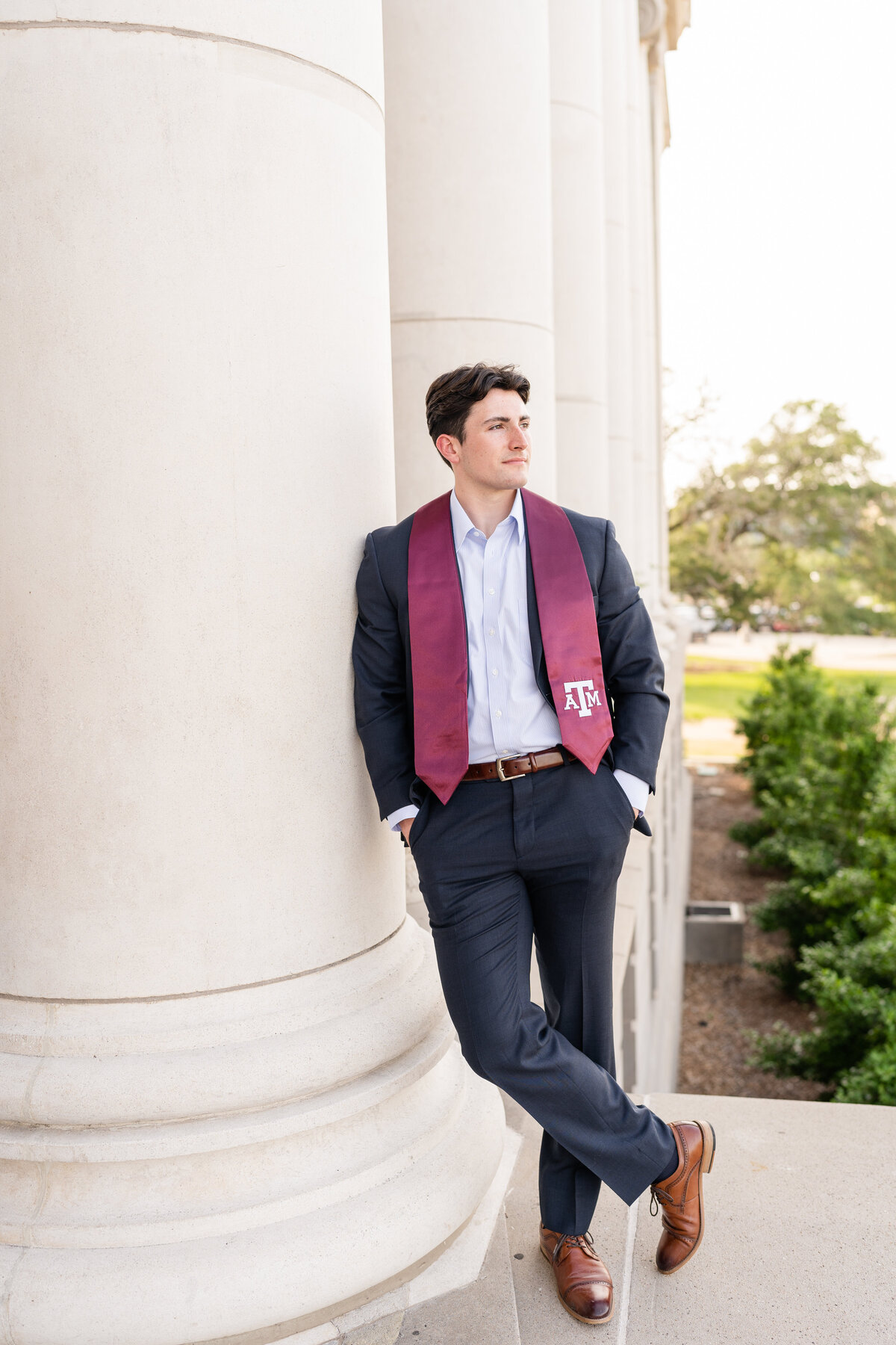 Texas A&M senior guy wearing a suit and Aggie stole with hands in pockets, leaning on a column and looking away at the Administration Building