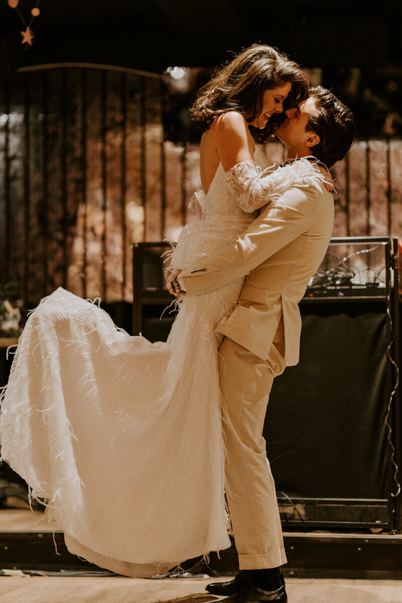 Marié en costume beige soulevant la mariée dans une tenue vintage. Décor industriel immortalisé par Laura Termeau photographie.