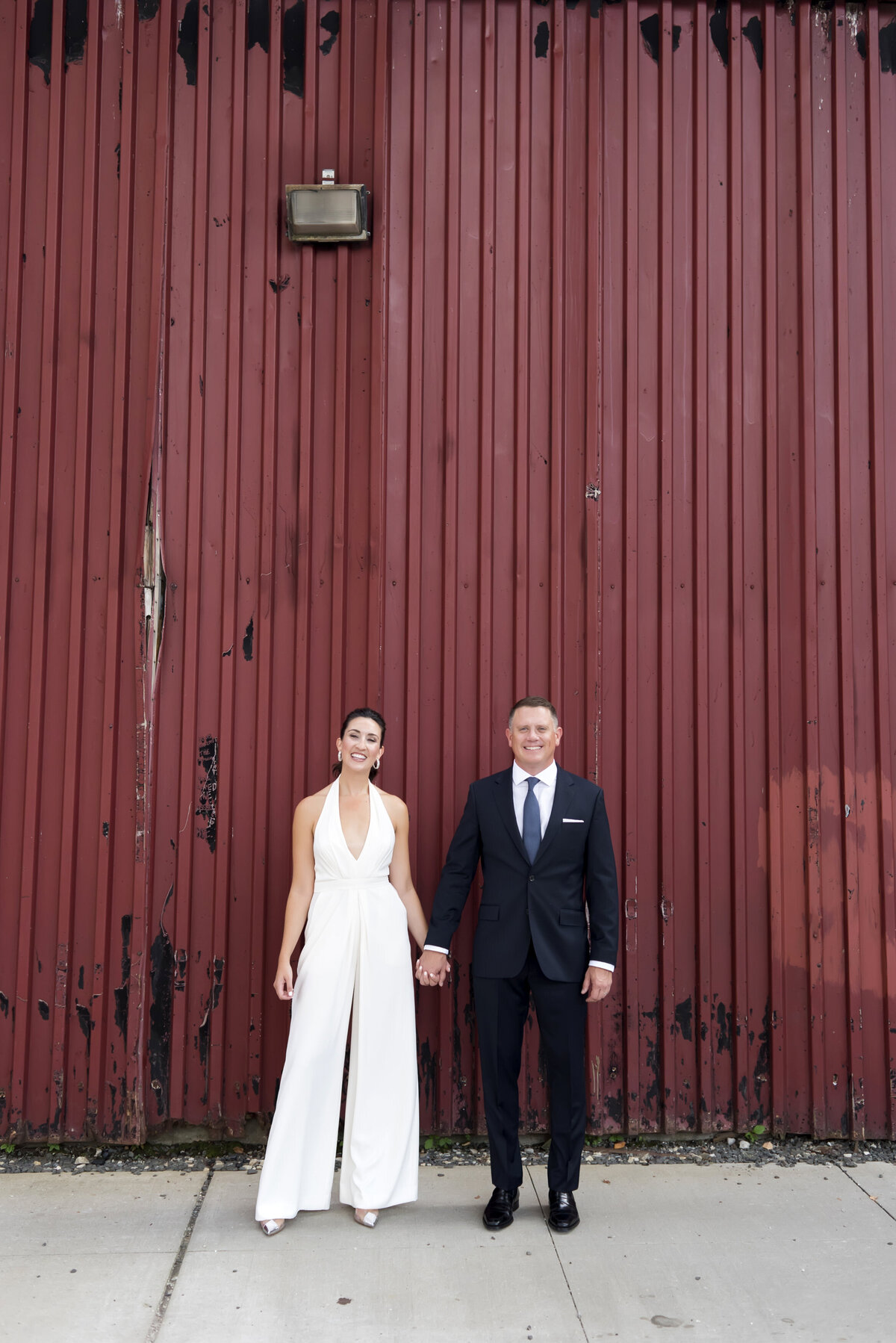 A wedding couple holding hands and standing outside a red building 