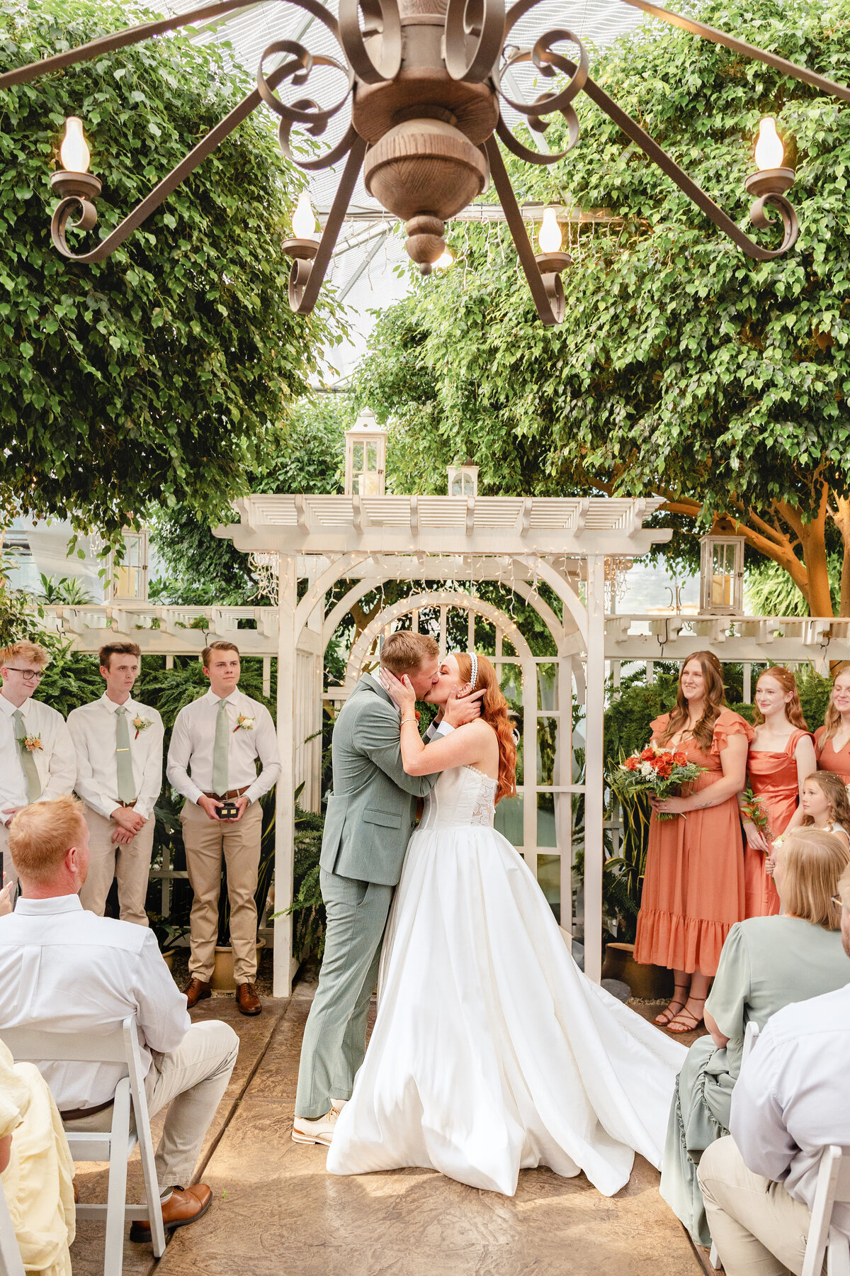 The bride and groom kiss at the altar, with their wedding party in the background, beneath an elegant white pergola adorned with fairy lights.