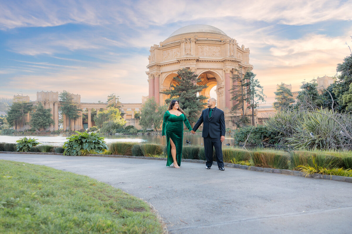 Engaged couple walk on a pathway in front of the Palace of Fine Arts in San Francisco. The man leads while the woman in a green dress follows while holding hands, smiling and looking at him.  Photo by sacarmento wedding photographer, philippe studio pro.