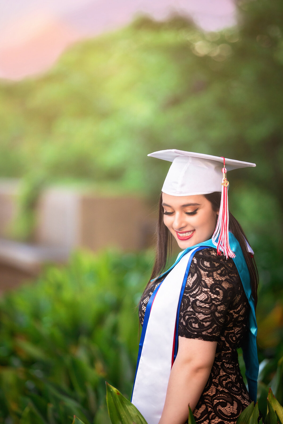Senior female with cap and tassel wearing graduation sashes.  She is looking sideways over her shoulder and wearing a black lace dress.  There is a pink sunset in the distance.