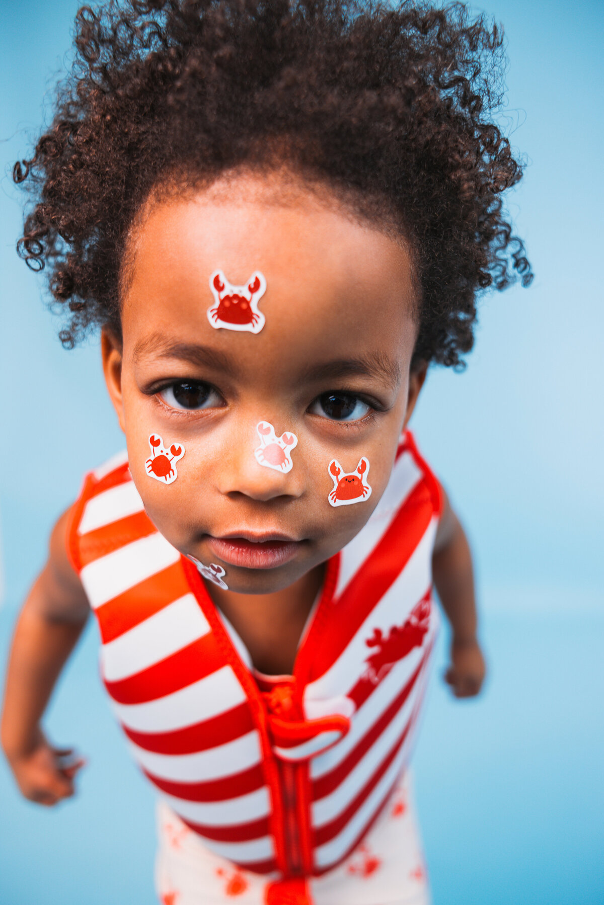 Child wearing a red-and-white striped Swim Essentials swim vest and crab-printed swim shorts, playfully decorated with crab stickers on their face, standing against a blue background.