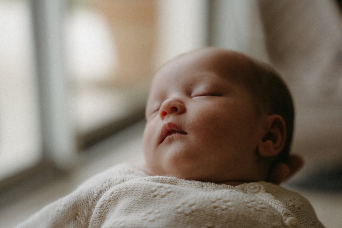 Bébé endormi, habillé de blanc, capturé en douceur près de la fenêtre et de la lumière naturelle. Séance naissance par Kamescop.