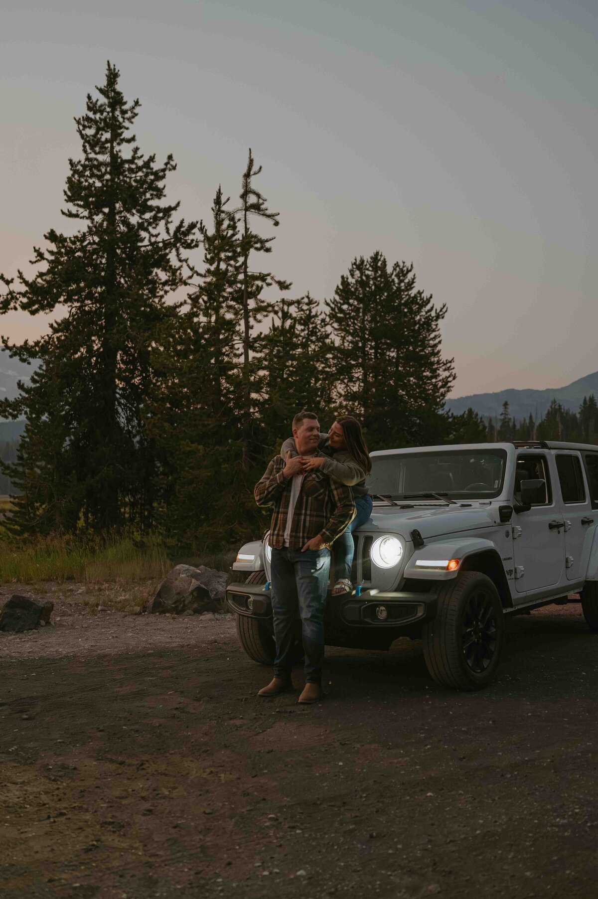 girl sitting on the hood of jeep hugging guy from behind with the headlights illuminating them