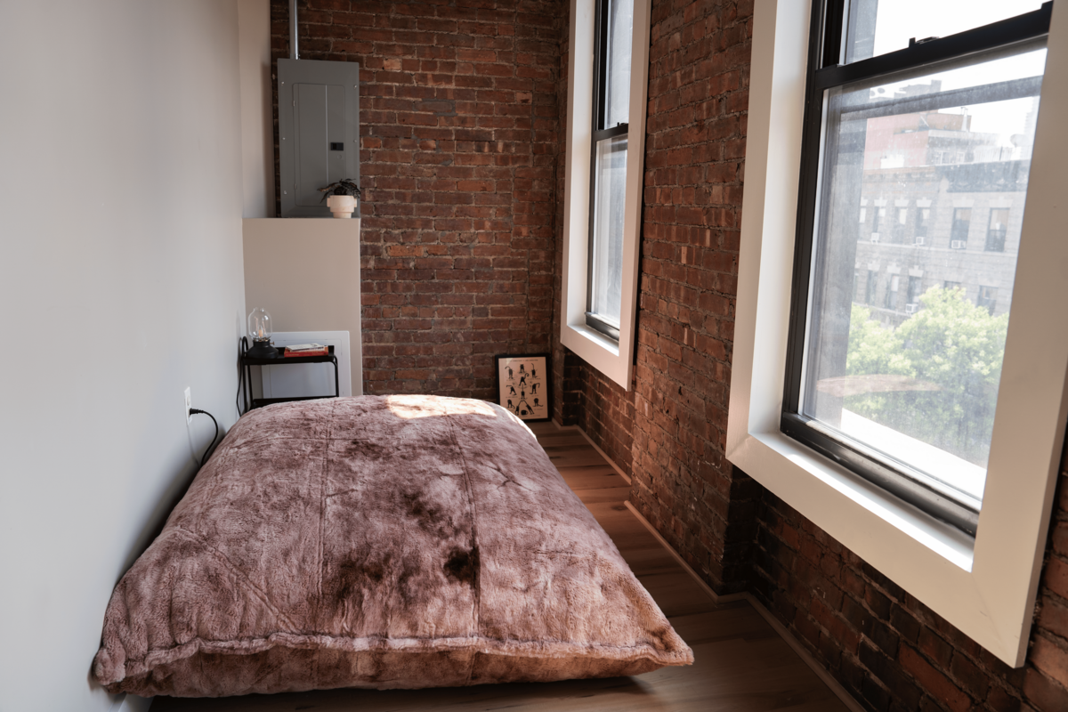 Cozy reading nook with a large cushion in a brick-walled corner, looking out through tall rectangular windows.