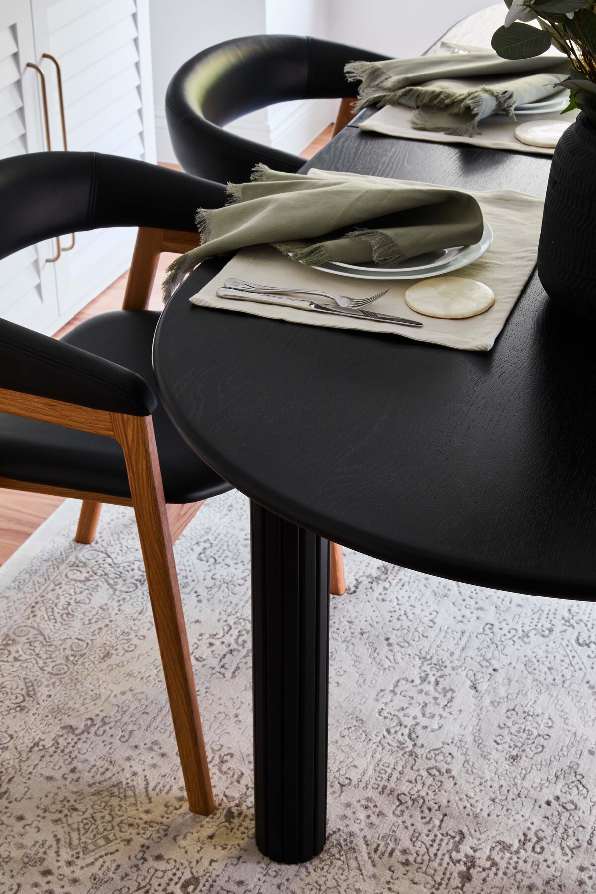 Black timber table set with sage napkins, linen placemats and ceramics beside sculptural oak dining chairs in a Williamstown home.