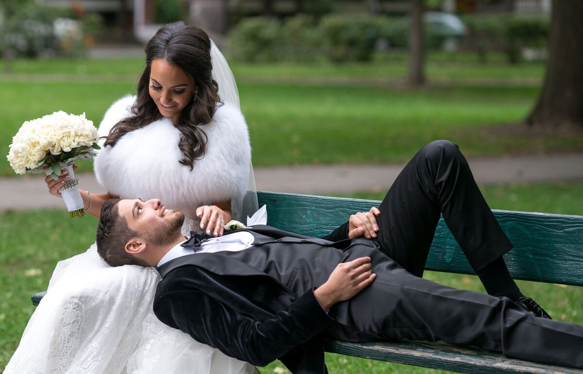 A tender scene of the groom lying down with his head resting on the bride's lap as they relax on a park bench. The serene outdoor setting and the couple's intimate pose capture a moment of quiet connection on their special day.