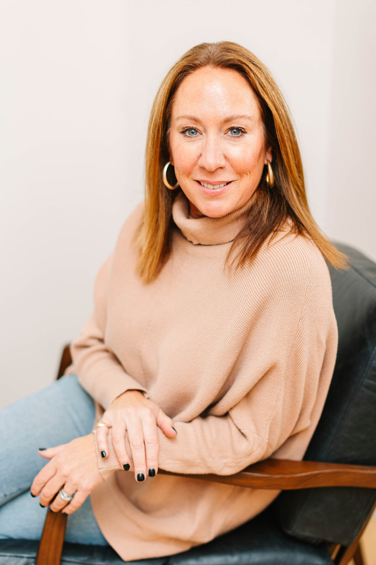A woman with straight, shoulder-length brown hair sits in a black leather chair, wearing a cozy camel-colored turtleneck sweater and light jeans. She smiles warmly, accessorized with gold hoop earrings and black nail polish, against a neutral background