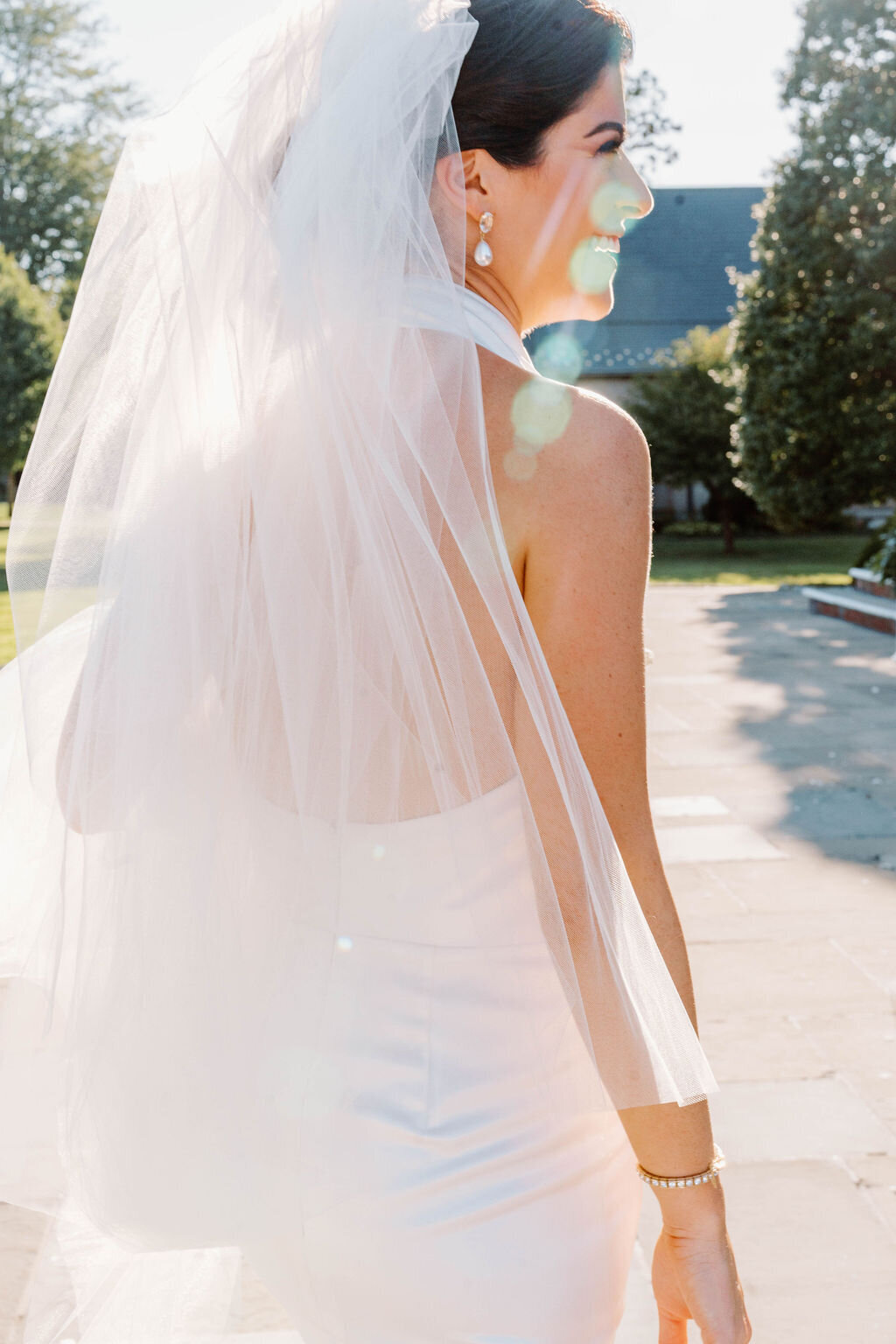 Wedding Photographer RI | Bride with a delicate, flowing veil stands outdoors in sunlight, creating a serene and joyful atmosphere. The background shows greenery and soft light flares.